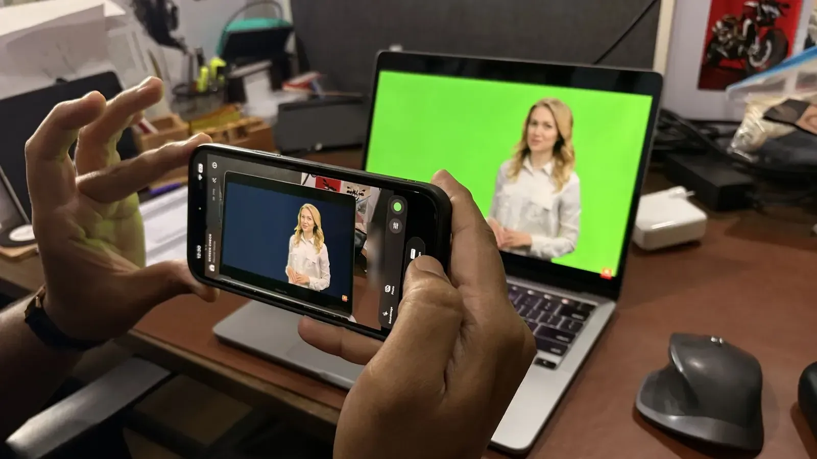 A phone running Screen Check held in front of a laptop playing green-screen footage. The phone shows the same shot already keyed onto a blue background.